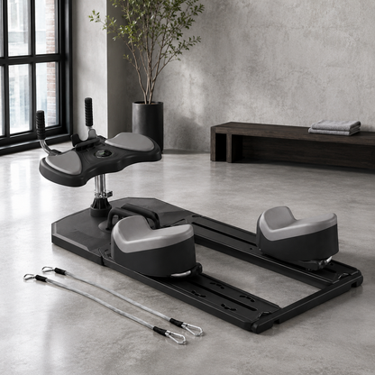 A black and gray seated leg exercise machine with two resistance bands placed on the floor, photographed in a modern minimalist room with concrete walls and a bench in the background.